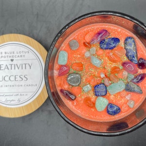 May include: A soy wax candle with embedded crystals and glitter. The candle is in a clear glass jar with a black lid. The label on the jar reads "Creativity Success Soy Wax Intention Candle".