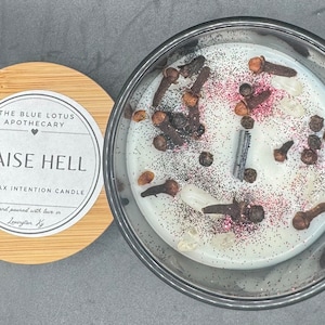 May include: A soy wax intention candle with a black lid and a wooden lid with a label that reads "The Blue Lotus Apothecary" and "Raise Hell" with a heart symbol. The candle is white and contains black peppercorns, cloves, and glitter.