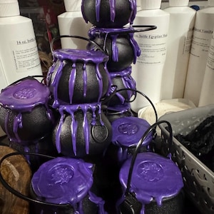May include: A collection of black cauldron-shaped candle holders with purple wax dripping down the sides. The candles are arranged in a pyramid shape and are surrounded by bottles of essential oils.