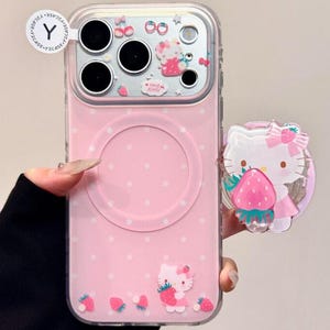 Puede incluir: Funda de teléfono rosa con diseño de Hello Kitty, con temática de fresas. La funda tiene un patrón de lunares e incluye un soporte a juego. El diseño incorpora fresas y el texto "Hello Kitty".