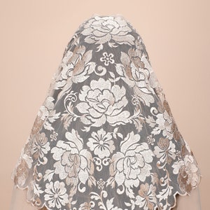May include: A white lace mantilla with a floral pattern in a beige colour. The lace is delicate and intricate, with a scalloped edge.