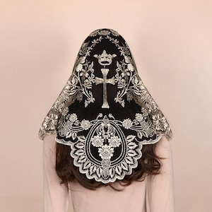 May include: Black lace head covering with intricate floral and cross embroidery. The veil features a crown design and a scalloped edge. The embroidery is in a contrasting light color.