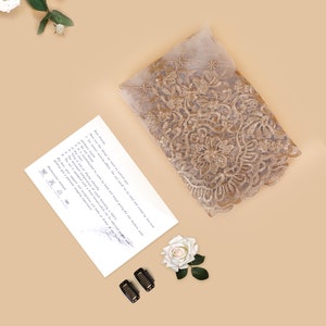 May include: A folded piece of beige lace fabric with gold embroidery. The fabric is decorated with floral patterns. The image also includes a white rose, a handwritten note, and two black hair clips.