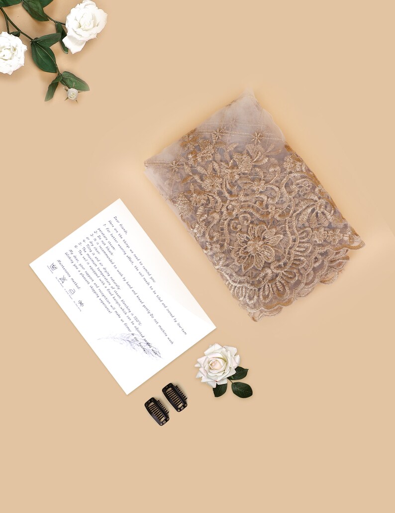 May include: A folded piece of beige lace fabric with gold embroidery. The fabric is decorated with floral patterns. The image also includes a white rose, a handwritten note, and two black hair clips.