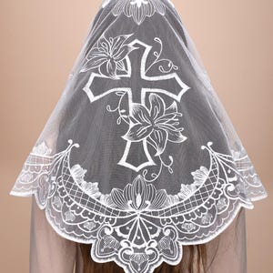May include: A white lace veil featuring a cross and floral pattern. The veil is worn over the head and shoulders, showcasing detailed lace work. The cross is central, with floral motifs surrounding it, creating an elegant design.