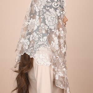 May include: A white lace mantilla with a floral pattern in a beige colour. The lace is delicate and intricate, with a scalloped edge.