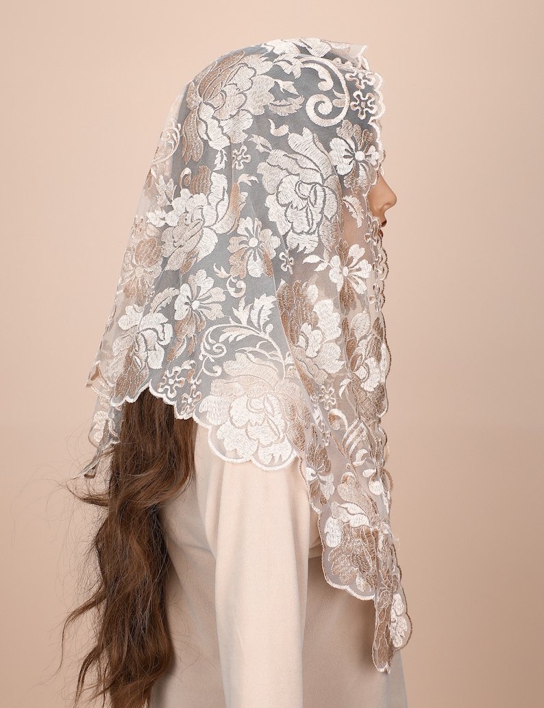 May include: A white lace mantilla with a floral pattern in a beige colour. The lace is delicate and intricate, with a scalloped edge.
