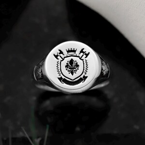 May include: A silver signet ring with a detailed crest design. The ring features a circular face with a central shield, crown, axes, and floral elements. The ring is set against a dark background, highlighting its polished finish.