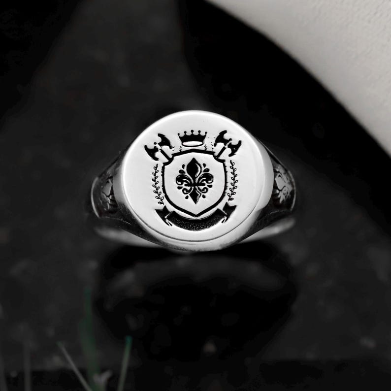 May include: A silver signet ring with a detailed crest design. The ring features a circular face with a central shield, crown, axes, and floral elements. The ring is set against a dark background, highlighting its polished finish.