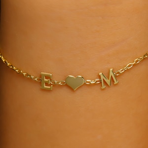May include: A gold chain bracelet with the letters "E" and "M" separated by a heart charm.