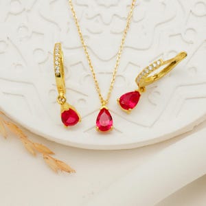 May include: A gold necklace and earrings set with a teardrop-shaped red gemstone pendant and matching earrings. The earrings are hoop style with a row of small clear stones.