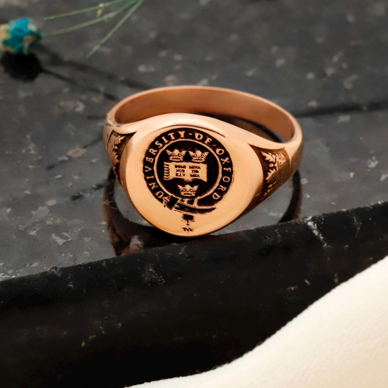 May include: A rose gold signet ring with the University of Oxford crest engraved on the oval face. The ring has a polished finish and features intricate detailing on the shoulders. The ring is displayed on a dark, textured surface.