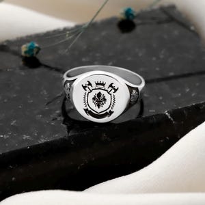 May include: A silver signet ring with a detailed crest design. The crest features a fleur-de-lis, axes, and a crown, all in black against a white background. The ring is set on a dark, textured surface.