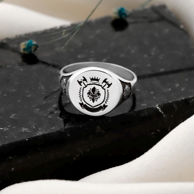 May include: A silver signet ring with a detailed crest design. The crest features a fleur-de-lis, axes, and a crown, all in black against a white background. The ring is set on a dark, textured surface.