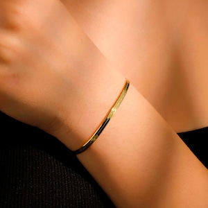 May include: A gold-toned bracelet with a simple, flat design. The bracelet is worn on a person's wrist.