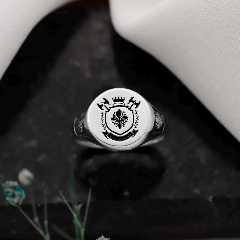 May include: A silver signet ring with a detailed crest design. The crest features a fleur-de-lis, crown, and other heraldic elements. The ring is set against a dark background, highlighting its polished finish.