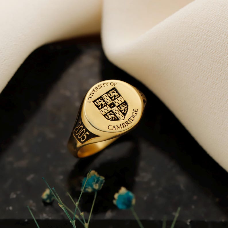 May include: A gold signet ring with the words "UNIVERSITY OF CAMBRIDGE" and the year "2005" engraved on it. The ring features a shield emblem with a detailed design. The ring is set against a dark background with a white fabric.