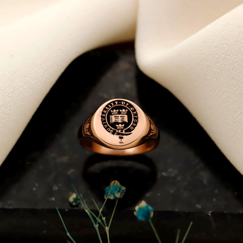 May include: Rose gold signet ring with the University of Oxford crest. The ring has a polished finish and a detailed engraving of the university seal. The ring is displayed on a dark surface with a white cloth in the background.