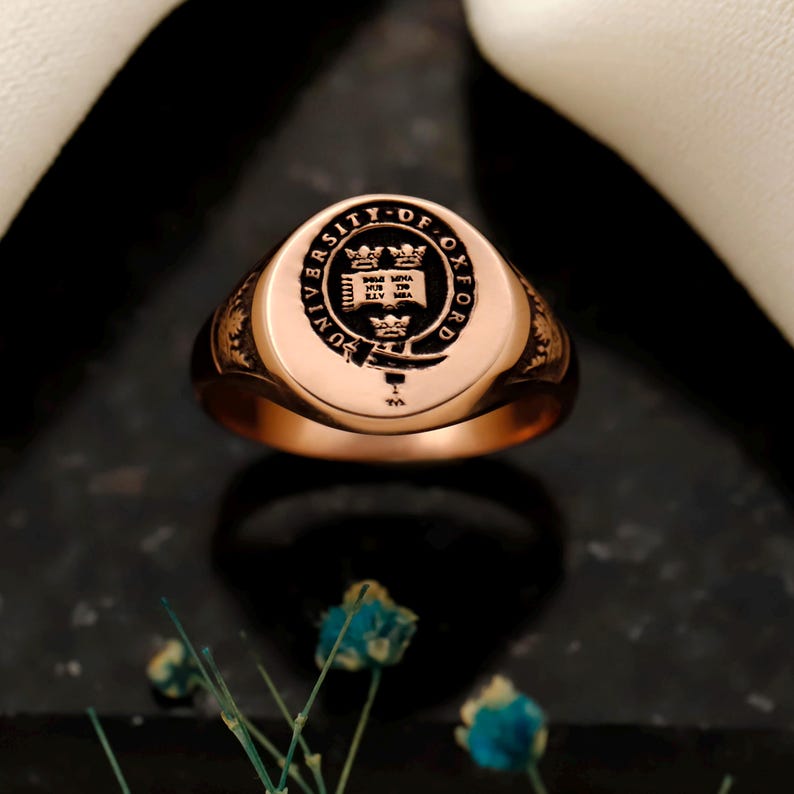 May include: A rose gold signet ring with the University of Oxford crest engraved on the face. The ring has a polished finish and a rounded, oval shape. The crest features a shield, crowns, and text around the border.