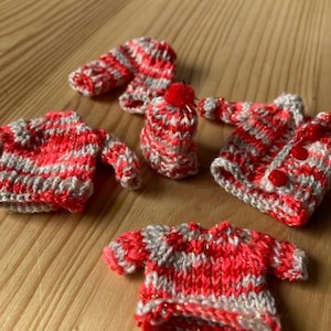 Könnte beinhalten: Eine Sammlung handgestrickter Miniaturkleidung in Rot, Grau und Gold. Die winzigen Kleidungsstücke, darunter Pullover, Mützen und Schals, sind auf einer Holzoberfläche angeordnet. Die Strickwaren zeigen detaillierte Muster und Texturen, mit Knöpfen an den Pullovern.