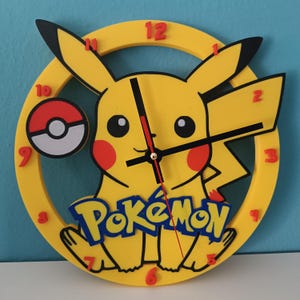 Può includere: Un orologio da parete giallo a tema Pikachu con orecchie nere, guance rosse e una Poké Ball. Il quadrante dell'orologio presenta la parola "Pokémon" in blu e giallo. I numeri delle ore sono rossi.