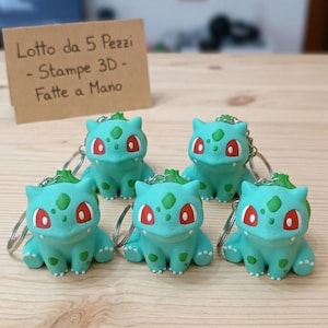Può includere: Cinque portachiavi a forma di creatura verde chiaro con occhi rossi e macchie verde scuro sul dorso. Ogni figura presenta una piccola struttura a bulbo sulla schiena ed è fissata a un anello in metallo argentato. Un cartoncino beige riporta il testo: Lotto da 5 Pezzi - Stampe 3D - Fatte a Mano.