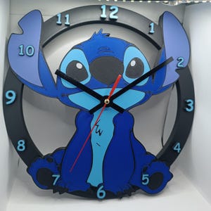 Peut inclure: Une horloge murale bleue et noire représentant le personnage Stitch. L'horloge a un cadre circulaire noir avec des chiffres et des aiguilles bleus. Le personnage est au centre, avec de grandes oreilles et une trotteuse rouge.
