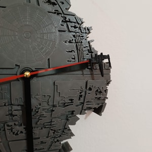 Puede incluir: Un reloj de pared gris impreso en 3D con la forma de la Estrella de la Muerte de Star Wars. El reloj tiene un segundero rojo, manecillas de minutos y horas negras y un centro dorado. El reloj está montado en una pared blanca.