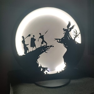 Può includere: Una lampada rotonda e illuminata con una scena silhouette. Il design nero ritagliato raffigura figure su un ponte, con un albero e una figura angelica a destra, su uno sfondo bianco brillante. La lampada ha una base nera.