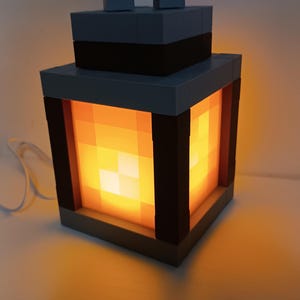 May include: A Minecraft-themed lantern made of gray and black blocks. The lantern emits a warm, orange glow from within, creating a pixelated light effect. The design includes a handle at the top and a square base. The lantern is plugged into a white cord.