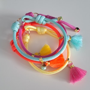 Könnte beinhalten: Vier farbige Armbänder aus Seil mit Quasten. Die Armbänder sind in Blautönen, Gelb, Rosa und Orange. Jedes Armband hat einen goldenen Akzent und eine Quaste.