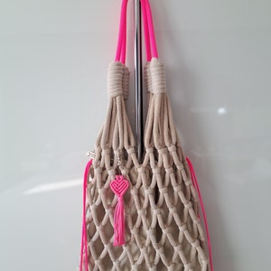 Puede incluir: Una bolsa de malla de crochet beige con asas de cuerda rosa y un borla rosa en forma de corazón. La bolsa está colgada de un gancho de metal.