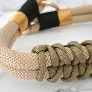 Könnte beinhalten: Nahaufnahme eines beige- und olivgrünen Paracord-Hundehalsbandes mit einer schwarzen und goldenen Metallschnalle.