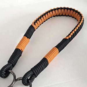 Può includere: Un cinturino in paracord nero e arancione con due anelli metallici. Il cinturino è intrecciato e presenta un motivo a colori contrastanti.