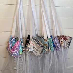 May include: A collection of reusable produce bags hanging from a decorative metal rack. Each bag features a different patterned fabric top, with a white mesh body. The fabrics include mermaid scales, floral, and text designs.