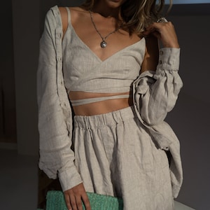 Puede incluir: Un top de lino beige con un diseño de envoltura y pantalones cortos de lino a juego. El top tiene un escote en V y un collar con un colgante plateado.