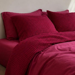 May include: A deep magenta bedding set featuring a waffle-textured duvet cover and matching pillowcases. The bed is dressed in solid, rich berry-toned linens, showcasing a prominent grid-like pattern on the top layer of the bedding ensemble.