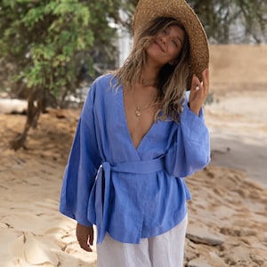 May include: A woman wearing a light blue linen wrap top with a wide, flowing neckline and long sleeves. She is wearing a straw hat and a gold necklace. The top is tied at the waist with a matching sash.