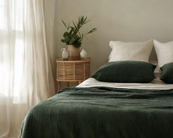 Puede incluir: Una funda nórdica de lino verde en una cama con fundas de almohada de lino blanco. La cama está decorada con dos almohadones de lino verde. Una pequeña mesita de noche de madera con una planta y elementos decorativos se encuentra en el fondo.