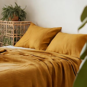 May include: A mustard yellow linen bedding set with two matching pillowcases. The bedding is arranged on a bed with a wooden side table and a potted plant.