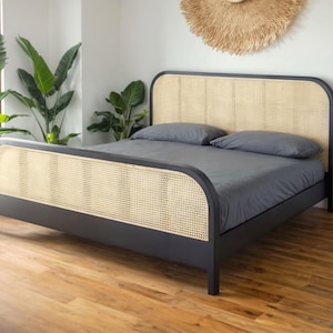 Puede incluir: Una estructura de cama con cabecero de ratán negro con un cabecero tejido y un colchón gris. La cama está en una habitación con suelo de madera y un gran tapiz de pared tejido.