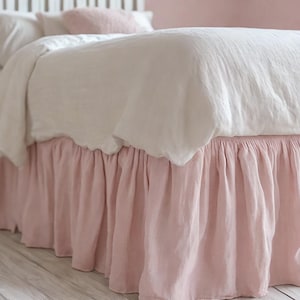 May include: A bed with white bedding and a light pink bed skirt. The bed skirt has a ruffled design. The bed is in a room with a white headboard and a white nightstand.