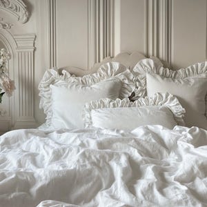 May include: White ruffled bedding with a white pillow and a white throw pillow on a bed with a white headboard.