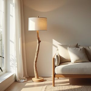 May include: A floor lamp with a natural wood trunk-style base and a beige lampshade. The lamp is next to a beige sofa with several pillows. A window with white curtains is on the left, and a potted plant is in the corner.