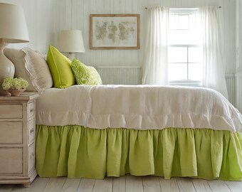 May include: A bed with a white duvet and a lime green valance. The bed is in a bright room with white walls, a window with white curtains, and a wooden framed picture on the wall. Two lamps sit on a bedside table.