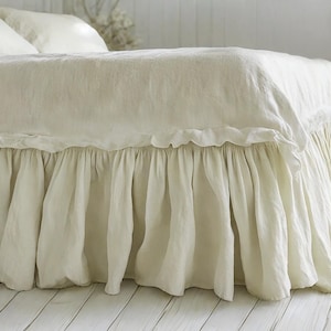 May include: Cream-colored bed linens with a ruffled bed skirt. The bedding includes a duvet cover and pillows, all in a soft, neutral tone. The bed skirt has a gathered, flowing design. The bed is set on a white wooden floor.