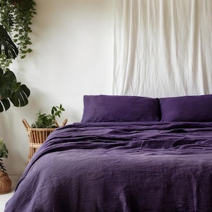 May include: A purple linen bedding set with two pillowcases and a duvet cover. The bedding is on a bed with a white headboard and a white curtain behind it.