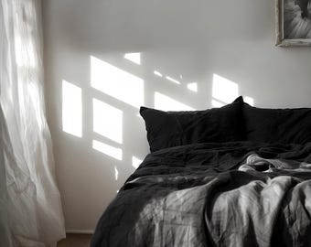 May include: A black linen bed with a white linen curtain in the background. The bed is made with a black duvet and two black pillows. Sunlight is shining through the window, casting a shadow on the wall.