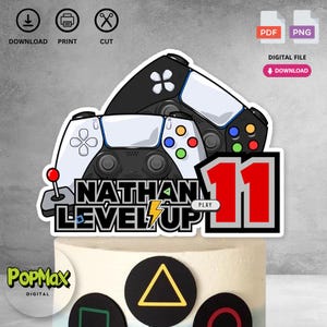 May include: A cake topper with two game controllers, one black and one white, with the text "NATHAN LEVEL UP" and the number "11". The design includes a lightning bolt and colourful buttons, ideal for a birthday celebration.