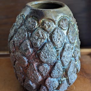 Pode incluir: Um vaso de cerâmica artesanal com uma superfície texturizada e orgânica. O vaso apresenta uma paleta de cores escuras e salpicadas com tons de castanho, cinza e toques de brilho metálico. O design e o acabamento únicos do vaso o tornam uma peça decorativa.
